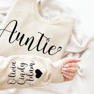 May include: A beige sweatshirt with the word "Auntie" in black cursive lettering on the front. The left sleeve has the names "Olivia, Cindy, Adam" in black lettering with a black heart.