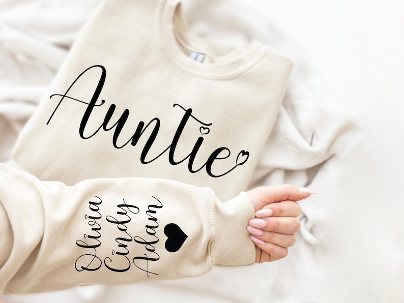May include: A beige sweatshirt with the word "Auntie" in black cursive lettering on the front. The left sleeve has the names "Olivia, Cindy, Adam" in black lettering with a black heart.