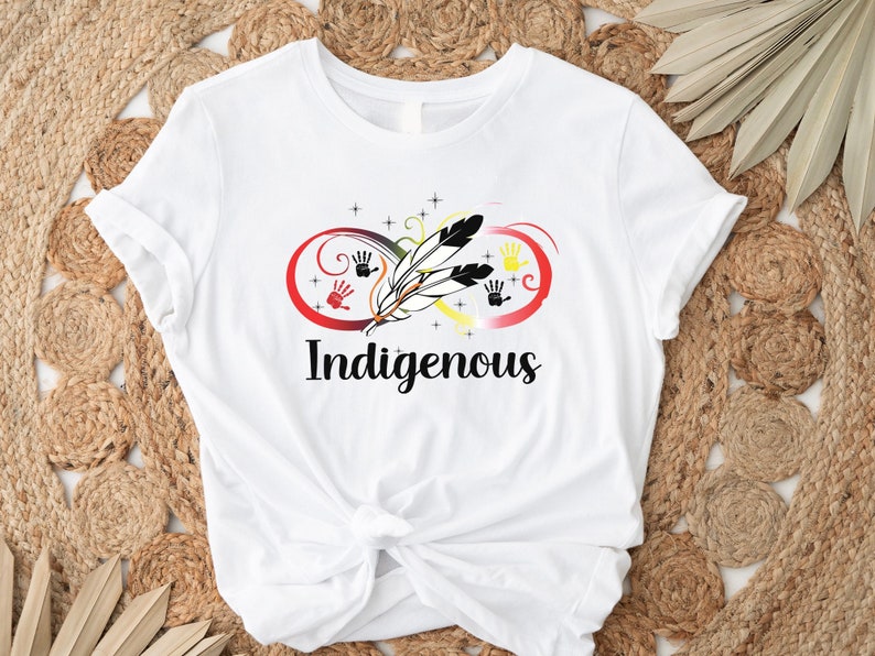 May include: White t-shirt featuring a graphic design of an infinity symbol with feathers and handprints in red, yellow, black, and white. The text "Indigenous" is printed below the graphic.
