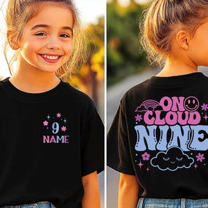 May include: Black t-shirt with a colorful design. The front features the number 9 and the word "NAME" in a pink and blue font. The back of the shirt says "ON CLOUD NINE" with a rainbow, smiley face, cloud, and flowers.