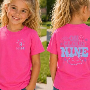 May include: A bright pink t-shirt with the text "9" and "N M" on the front. The back of the shirt features the phrase "On Cloud Nine" with a rainbow, smiley face, stars, and a cloud graphic. The shirt is short-sleeved.