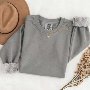 K&ouml;nnte beinhalten: Ein graues Sweatshirt mit Rundhalsausschnitt und Comfort Colors Etikett, erg&auml;nzt durch eine goldfarbene Kette mit kleinem Sternanh&auml;nger. Das Sweatshirt hat lange &Auml;rmel mit gerippten B&uuml;ndchen und liegt neben einem beigen Hut und Trockenblumen.