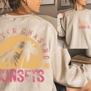 May include: A beige sweatshirt with a graphic print of a sunset and palm trees. The text "FOREVER CHASING SUNSETS" is printed on the back of the sweatshirt.