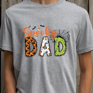 May include: A gray t-shirt with the words "Spooky DAD" in orange and various Halloween-themed designs. The letters are decorated with bats, pumpkins, and a spider web. The shirt is made of a soft material.