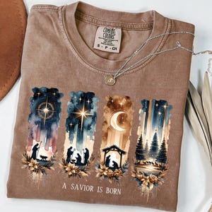 May include: Brown Comfort Colors t-shirt featuring a watercolor design of the Nativity scene. The design includes a star, moon, and silhouettes of figures. The text "A Savior Is Born" is printed at the bottom. A silver necklace is draped across the shirt.