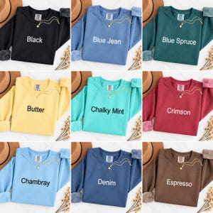 K&ouml;nnte beinhalten: Ein Raster aus neun Rundhals-Sweatshirts in verschiedenen Farben, darunter Schwarz, Blue Jean, Blue Spruce, Butter, Chalky Mint, Crimson, Chambray, Denim und Espresso. Jedes Sweatshirt hat eine kleine goldene Sternhalskette und ein Markenetikett.