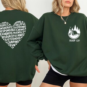May include: Dark green sweatshirt with white text and graphics. The back features a heart-shaped design with words like "Camping", "Kindness", and "Fun". The front has a camping scene and the text "Troop 123".