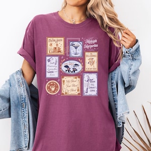 May include: A maroon t-shirt featuring a graphic print of multiple rectangular postage stamp designs. The stamps include illustrations of a house, a margarita, a witch hat, and text such as Midnight Margaritas, The Belladonna, and My Blood Your Blood Our Blood.