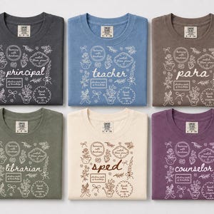 May include: Six folded crew neck t-shirts in charcoal, blue, brown, olive green, cream, and purple. Each shirt features a white or brown graphic print of floral doodles, bows, and text including principal, teacher, para, librarian, sped, and counselor.