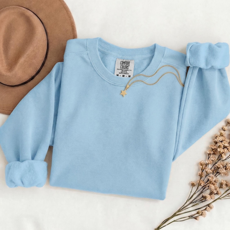 K&ouml;nnte beinhalten: Ein hellblaues Sweatshirt mit Rundhalsausschnitt und einem Comfort Colors Markenetikett am inneren Halsausschnitt. Eine goldfarbene Halskette mit einem kleinen sternf&ouml;rmigen Anh&auml;nger liegt auf dem Stoff. Das Sweatshirt hat lange &Auml;rmel mit Rippb&uuml;ndchen.