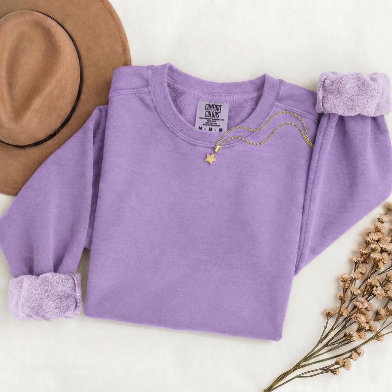 K&ouml;nnte beinhalten: Ein lavendelfarbenes Sweatshirt mit Rundhalsausschnitt, langen &Auml;rmeln und gerippten B&uuml;ndchen, das am inneren Halsausschnitt ein Etikett der Marke Comfort Colors aufweist. Eine zarte goldfarbene Kette mit einem kleinen sternf&ouml;rmigen Anh&auml;nger liegt darauf.