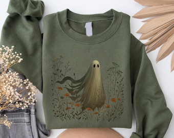 May include: An olive green crewneck sweatshirt featuring a central graphic of a pale, translucent ghost floating amidst a field of wildflowers. The illustration includes delicate stems, leaves, and small orange blossoms surrounding the ghost figure.