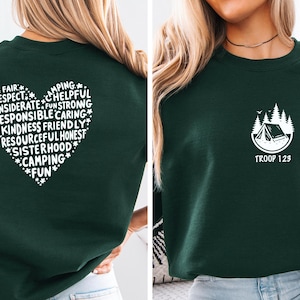 May include: Dark green sweatshirt with a white heart-shaped graphic on the back, filled with words like "respect" and "kindness." The front features a camping-themed logo with the text "TROOP 123."