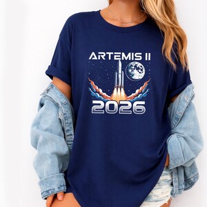 May include: Navy blue t-shirt featuring a graphic of a rocket launching into space, with the moon and stars in the background. The text "ARTEMIS II" and "2026" are printed in white. The shirt is worn with a denim jacket.
