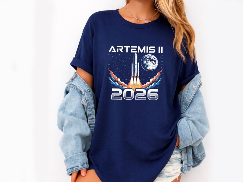 May include: Navy blue t-shirt featuring a graphic of a rocket launching into space, with the moon and stars in the background. The text "ARTEMIS II" and "2026" are printed in white. The shirt is worn with a denim jacket.