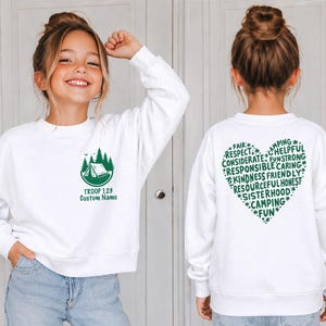 May include: White sweatshirt with a green camping graphic and text "TROOP 123 Custom Name" on the front. The back features a green heart-shaped design with words like "FAIR", "RESPECT", and "CAMPING".