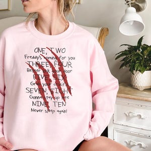 May include: A light pink sweatshirt with the text "ONE, TWO Freddy's coming for you THREE, FOUR Better lock your door FIVE SIX Grab your crucifix SEVEN, EIGHT Gonna stay up late NINE TEN Never sleep again!" and red claw marks.