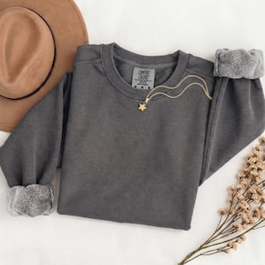 K&ouml;nnte beinhalten: Ein anthrazitfarbenes Sweatshirt mit Rundhalsausschnitt, langen &Auml;rmeln und gerippten B&uuml;ndchen. Das Innenetikett zeigt Comfort Colors, Gr&ouml;&szlig;e M. Eine goldfarbene Kette mit einem kleinen Sternanh&auml;nger liegt am Halsausschnitt.