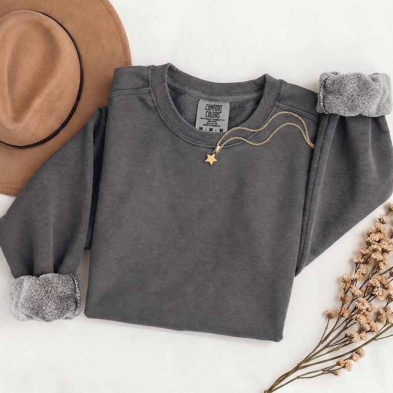 K&ouml;nnte beinhalten: Ein anthrazitfarbenes Sweatshirt mit Rundhalsausschnitt, langen &Auml;rmeln und gerippten B&uuml;ndchen. Das Innenetikett zeigt Comfort Colors, Gr&ouml;&szlig;e M. Eine goldfarbene Kette mit einem kleinen Sternanh&auml;nger liegt am Halsausschnitt.