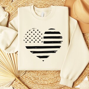 May include: A cream-coloured sweatshirt with a black and white distressed American flag heart design.
