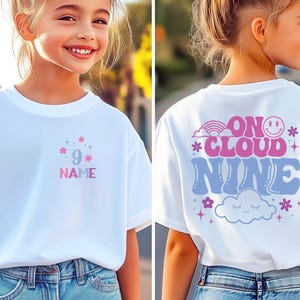May include: White t-shirt with a colorful design. The front features the number 9, stars, and the word "NAME" in pink and blue. The back says "ON CLOUD NINE" with a rainbow, smiley face, and cloud graphic.