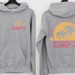May include: Gray hooded sweatshirt with a graphic of a sunset and palm trees on the back. The text "FOREVER CHASING SUNSETS" is printed in pink and yellow on the back. The front of the sweatshirt has a smaller graphic of a sunset with the text "SUNSETS" printed in pink and yellow.