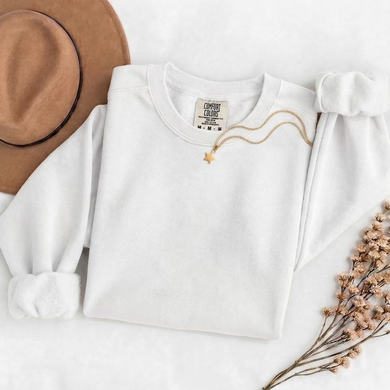 K&ouml;nnte beinhalten: Ein schlichtes wei&szlig;es Langarm-Sweatshirt mit Rundhalsausschnitt und einem Comfort Colors Markenetikett am inneren Halsausschnitt. Eine d&uuml;nne goldfarbene Kette mit einem kleinen sternf&ouml;rmigen Anh&auml;nger liegt auf dem Stoff. Ein brauner Filzhut liegt daneben.