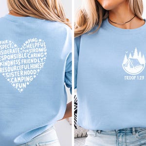 May include: Light blue sweatshirt with a white heart-shaped design on the back, filled with words like 'Respect' and 'Camping'. The front features a white camping scene with the text 'TROOP 123'.
