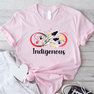 May include: Pink t-shirt featuring a graphic design of an infinity symbol with feathers and handprints in red, yellow, and black. The text "Indigenous" is printed below the graphic.