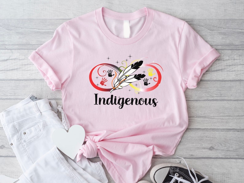 May include: Pink t-shirt featuring a graphic design of an infinity symbol with feathers and handprints in red, yellow, and black. The text "Indigenous" is printed below the graphic.