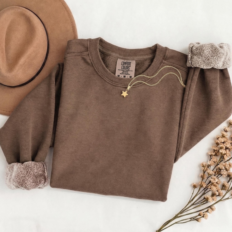 K&ouml;nnte beinhalten: Ein braunes Sweatshirt mit Rundhalsausschnitt und Comfort Colors-Etikett, ausgestattet mit langen &Auml;rmeln und gerippten B&uuml;ndchen. Eine goldfarbene Kette mit einem kleinen Sternanh&auml;nger liegt am Ausschnitt. Das Sweatshirt wird zusammen mit einem braunen Hut gezeigt.