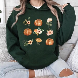 Puede incluir: Sudadera con capucha verde oscuro con un dise&ntilde;o de calabaza y flores. El dise&ntilde;o incluye calabazas naranjas y varias flores en tonos melocot&oacute;n, blanco y verde. La sudadera tiene un bolsillo delantero y una capucha.