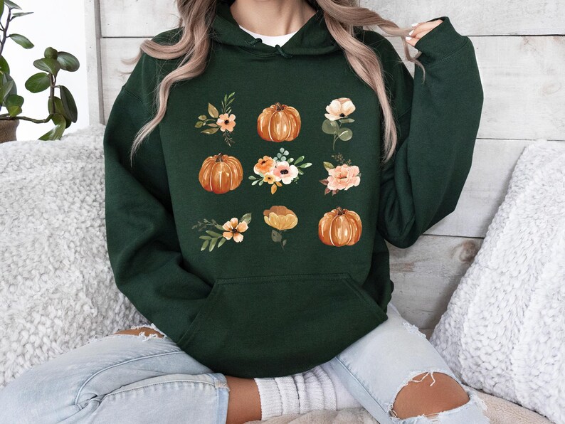 Puede incluir: Sudadera con capucha verde oscuro con un dise&ntilde;o de calabaza y flores. El dise&ntilde;o incluye calabazas naranjas y varias flores en tonos melocot&oacute;n, blanco y verde. La sudadera tiene un bolsillo delantero y una capucha.