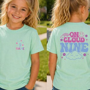 May include: A light green t-shirt with the text "9" and "NAME" in a pocket design. The back of the shirt has the words "ON CLOUD NINE" with a rainbow, smiley face, and cloud graphic.