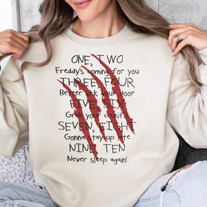 May include: Cream-colored sweatshirt with the text "ONE, TWO Freddy's coming for you... Never sleep again!" in black and red. The design includes red claw marks. A casual, horror-themed apparel item.