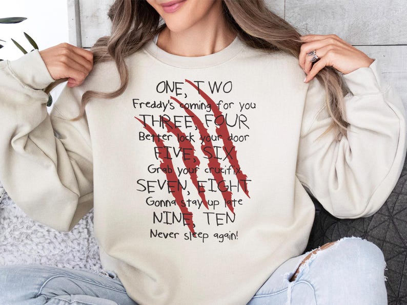 May include: Cream-colored sweatshirt with the text "ONE, TWO Freddy's coming for you... Never sleep again!" in black and red. The design includes red claw marks. A casual, horror-themed apparel item.