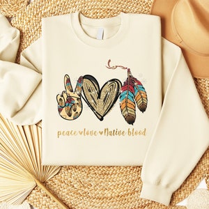 May include: A beige sweatshirt with a peace sign, heart, and feathers design. The text "peace love Native blood" is printed in gold below the design.