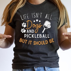 May include: A dark grey t-shirt with the text "Life isn't all dogs and pickleball but it should be" in white. The text is surrounded by a white paw print, a white pickleball paddle and ball, and five white stars.