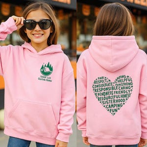 May include: A light pink hooded sweatshirt with a front pocket and a green graphic of a tent and trees. The text "TROOP 128 Custom Name" is below the graphic. The back of the sweatshirt has a heart-shaped design with words like "Fair", "Respect", and "Camping".