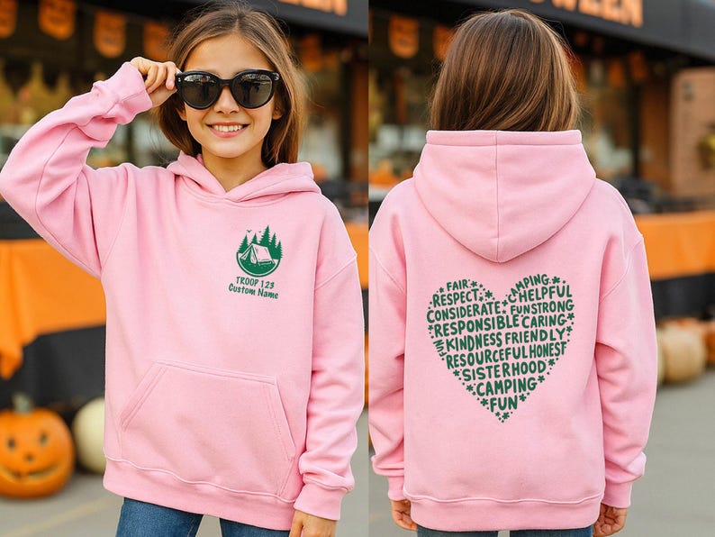 May include: A light pink hooded sweatshirt with a front pocket and a green graphic of a tent and trees. The text "TROOP 128 Custom Name" is below the graphic. The back of the sweatshirt has a heart-shaped design with words like "Fair", "Respect", and "Camping".