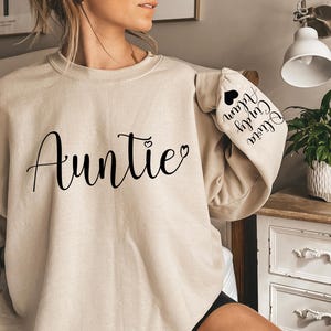 May include: A beige sweatshirt with the word "Auntie" printed on the front in black cursive lettering. The left sleeve has the text "Olivia, Colby, Alan" printed in black cursive lettering.