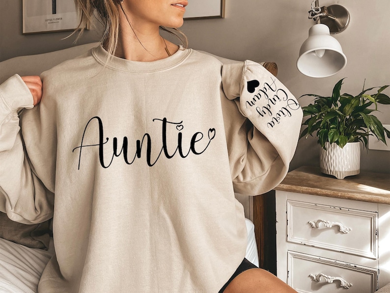 May include: A beige sweatshirt with the word "Auntie" printed on the front in black cursive lettering. The left sleeve has the text "Olivia, Colby, Alan" printed in black cursive lettering.