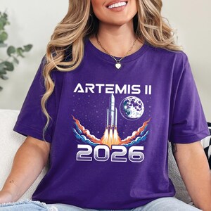 May include: Purple t-shirt featuring a graphic of a rocket launching into space, with the text "ARTEMIS II" above and "2026" below. The design includes a moon and stars. The shirt is a crew neck.