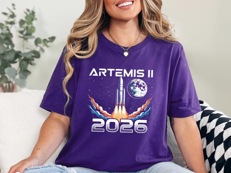May include: Purple t-shirt featuring a graphic of a rocket launching into space, with the text "ARTEMIS II" above and "2026" below. The design includes a moon and stars. The shirt is a crew neck.