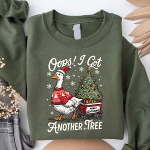 May include: Olive green sweatshirt with a graphic of a goose wearing a Santa hat and a red sweater, pulling a cart with a Christmas tree. Text reads "Oops! I Got Another Tree" and "Merry Goosemas".