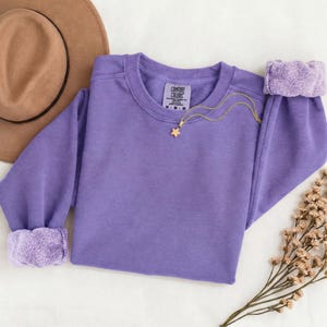 K&ouml;nnte beinhalten: Ein einfarbiges, violettes Langarm-Sweatshirt mit Rundhalsausschnitt und einem Comfort Colors-Markenetikett am inneren Halsausschnitt. Eine zarte goldfarbene Kette mit einem kleinen sternf&ouml;rmigen Anh&auml;nger liegt auf der Vorderseite.
