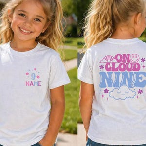 May include: White t-shirt with a colorful design. The front features the number 9 with the word "NAME" below it. The back of the shirt says "ON CLOUD NINE" with a rainbow, cloud, and flower graphics. A fun, casual tee.