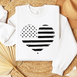 May include: White sweatshirt featuring a black and white heart-shaped design of the American flag. The flag has a distressed, grunge effect.