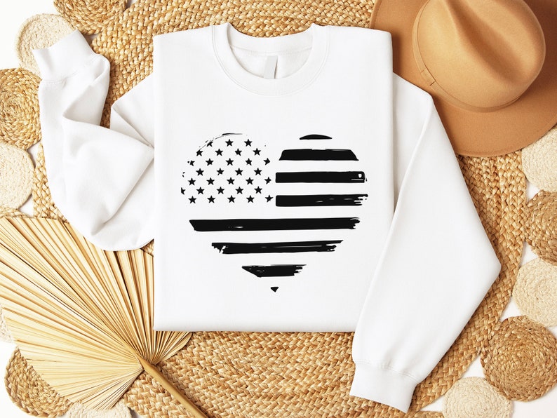 May include: White sweatshirt featuring a black and white heart-shaped design of the American flag. The flag has a distressed, grunge effect.