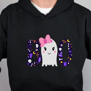May include: A black hooded sweatshirt featuring a graphic print of the word BOO. The letters B and O are filled with a dark purple pattern of skulls, bones, and cats. A white ghost with a pink bow replaces the letter O in the center.